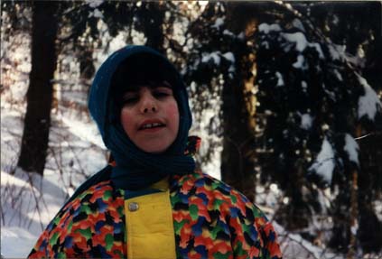 Enfant avec bonnet et écharpe bleue en anorak multicolore devant sapins enneigés.