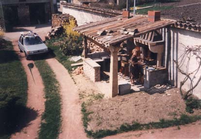 Femme sous un abri de jardin avec barbecue devant un chemin de terre et voiture.