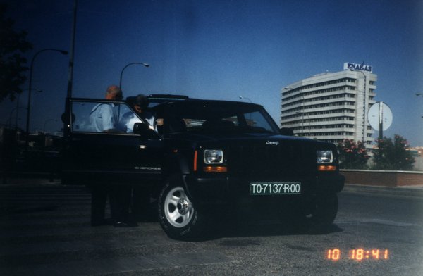Voiture noire de type 4X4 avec deux personnes debout dans l’embrasure de la portière. Date et heure en impression.
