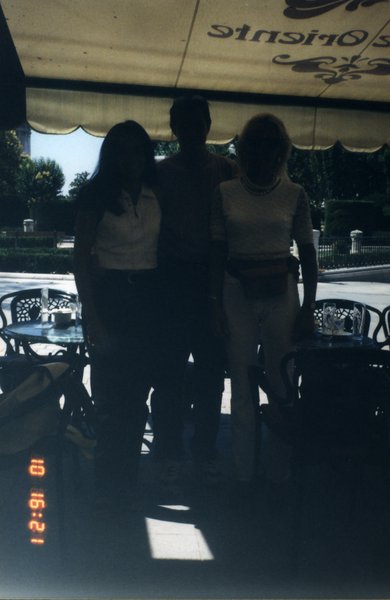 Homme debout entouré de deux femmes sous l’auvent d’une terrasse de café.
