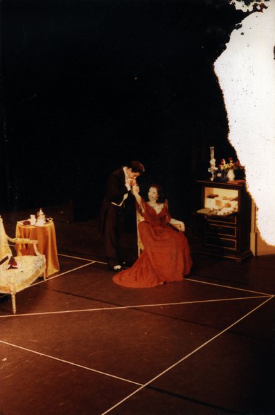Plateau de théâtre avec scène de baise-main entre un homme en frac et une femme en robe longue rouge décolletée assise devant une coiffeuse. Image avec tache blanche.