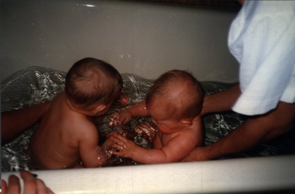 Deux bébés nus dans l’eau d’une baignoire, bras en avant-plan.