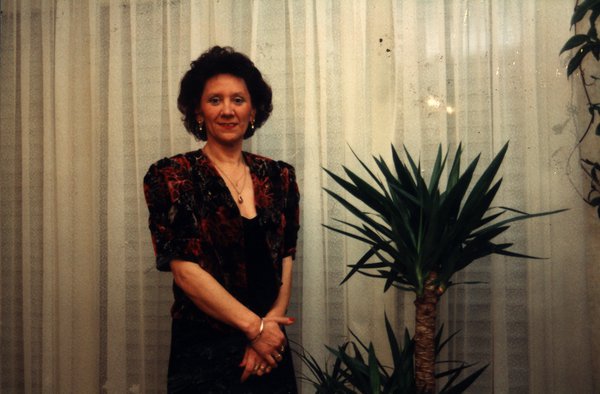 Femme brune élégante debout près d’une plante verte devant une fenêtre avec voilages blancs.