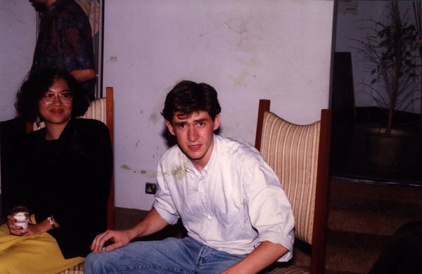 Jeune homme assis en jean et chemise blanche sur un siège à dossier rayé, à ses côté une femme, un gobelet à la main. Image avec salissures.