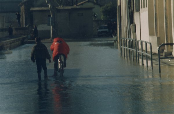 Personne en anorak rouge sur un vélo et une autre debout avec des bottes en caoutchouc dans une rue inondée par la pluie.