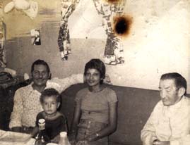Femme assise sur une banquette entourée de deux hommes et d’un enfant devant une table avec bouteilles. Image avec traces de brûlure. Noir et blanc.