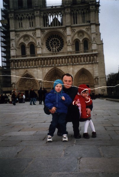 Homme accroupi devant la cathédrale Notre-Dame à Paris avec dans les bras un petit garçon en anorak et bonnet bleu et une petite fille en manteau rouge. Image avec déchirure, reconstituée.