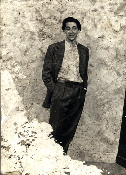 Homme brun à veste sombre et chemise blanche debout devant un mur avec les mains dans les poches de son pantalon. Image abîmée. Noir et blanc.