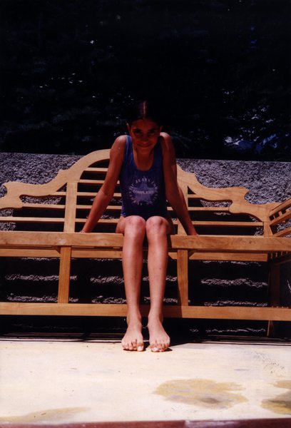 Petite fille en maillot une pièce bleu assise en plein air sur un banc en bois ouvragé.