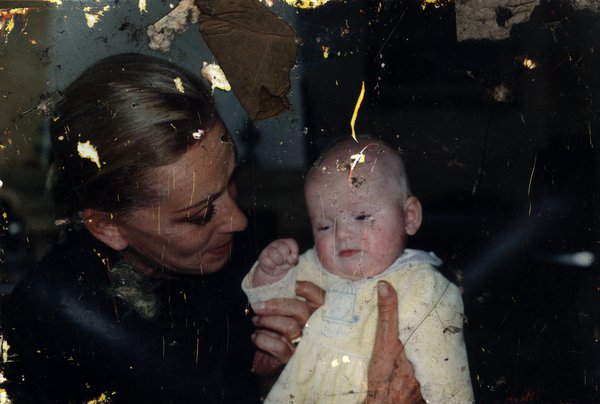 Femme blonde cheveux tirés avec un bébé en grenouillère jaune pâle. Image abîmée.
