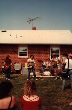 Concert avec guitaristes et batterie devant la façade d’un pavillon en briques rouges, femmes de dos en avant-plan.