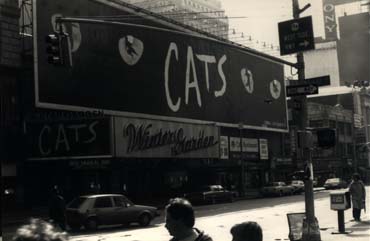 Panneau avec affichage pour : « Cats » dans une rue avec passants et voitures. Noir et blanc.
