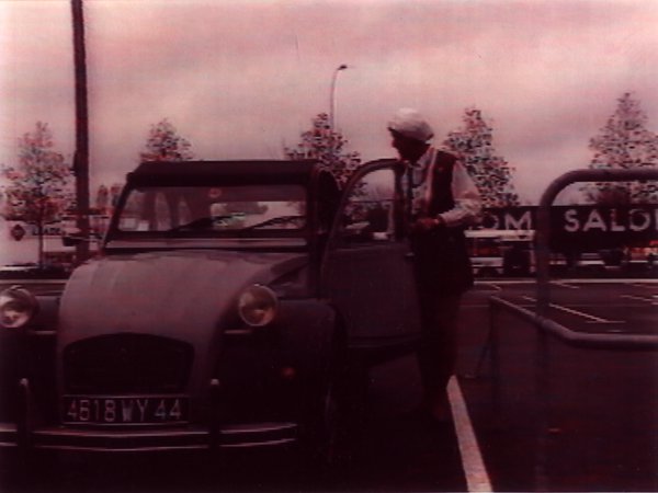 Citroën 2 CV et femme avec cheveux blancs ? debout portière-avant ouverte.
