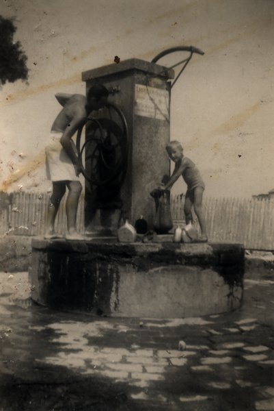 Un enfant blond et un adolescent debouts sur la margelle d’une fontaine à manivelle. Noir et blanc.