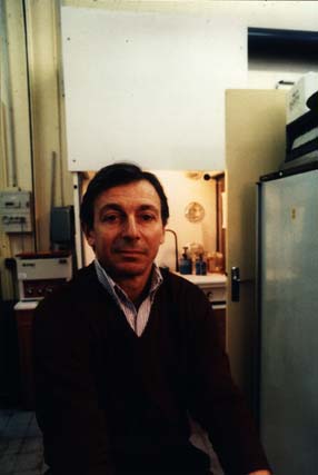 Dark-haired man sitting facing front, door and cupboard in the background.