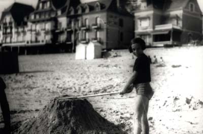 Une petite fille tasse à la pelle un tas de sable sur une plage, cabine de bain et maisons en arrière-plan. Noir et blanc.