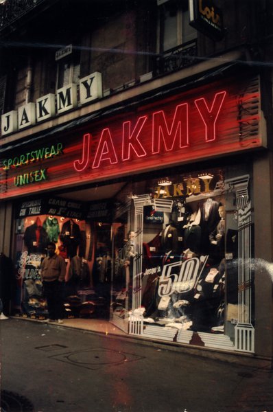 Devanture de magasin de vêtements avec enseigne lumineuse « Jakmy » de nuit.ᴬᴵ