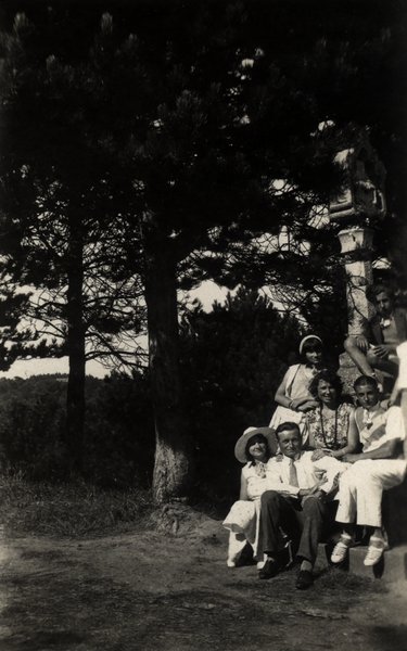 Groupe de personnes assises près d'un monument en pierre dans les pins. Noir et blanc.ᴬᴵ