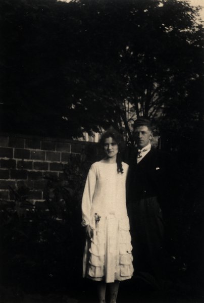 Jeune femme et homme debout dans un jardin, tenue habillée. Noir et blanc.ᴬᴵ