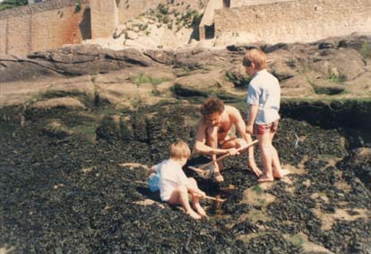 Homme à lunettes en maillot de bain accroupi dans les rochers avec une épuisette accompagné de deux enfants.