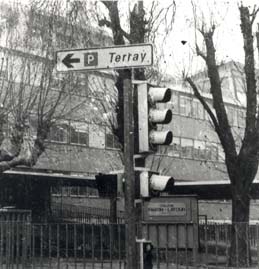 Feux de signalisation avec panneau de direction pour un parking devant grille et façade d’immeuble. Noir et blanc.