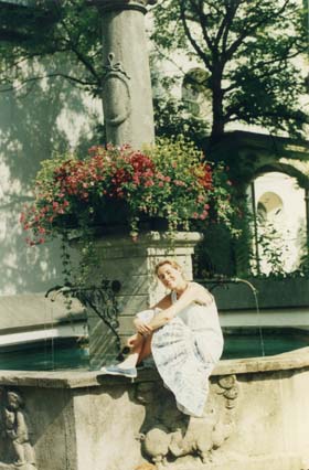 Jeune femme blonde souriante assise sur la margelle d’une fontaine ancienne.