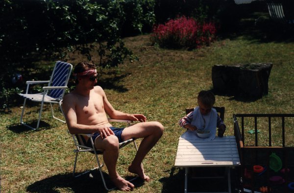 Homme torse nu assis dans un jardin près d'un enfant à table.ᴬᴵ