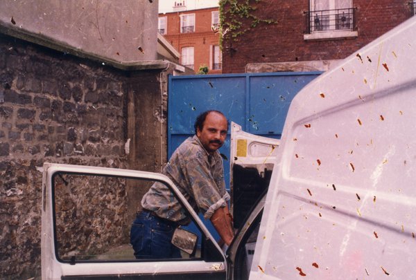 Homme moustachu en bras de chemise devant le capot ouvert d’une fourgonnette blanche. Image abîmée.