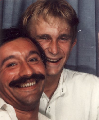 Visages d’un homme blond et d’un homme brun moustachu riants côte à côte.