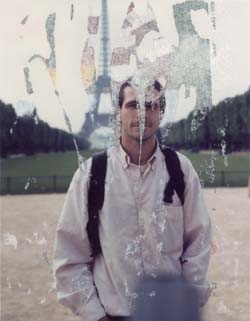 Buste d’homme brun en anorak blanc et sac à dos devant l’esplanade des Invalides et la Tour Eiffel. Image grattée, très abîmée.