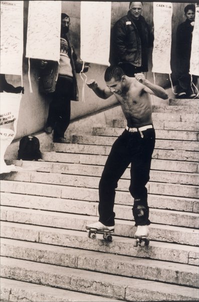 Descente en roller d’un escalier par un homme brun torse nu devant des manifestants à banderoles. Noir et blanc.