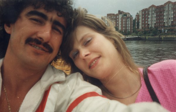Homme brun moustachu et femme blonde, leurs visages côte à côte, plan d’eau et immeubles en arrière-plan.