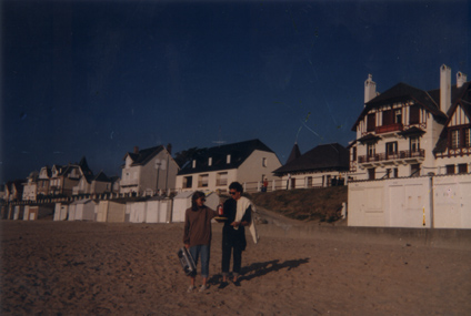 Jeune femme et jeune homme en lunettes noires sur une plage normande avec cabines de bains blanches.