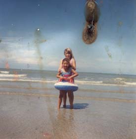 Une petite fille blonde et une autre avec une bouée bleue sur le sable devant la mer.