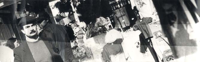 Homme brun à moustache devant une échoppe de gants et de vêtements dans la rue. Noir et blanc.