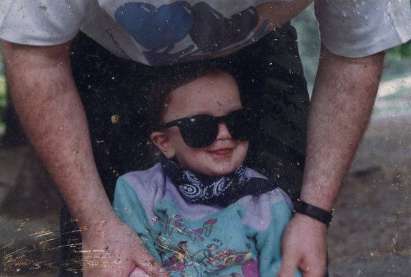 Homme penché bras nus sur un enfant avec des lunettes de soleil et un bandana bleu autour du cou.