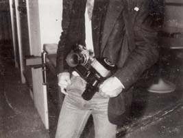 Buste d’homme en veste noir avec un appareil photo et flash en bandoulière, les mains dans les poches de son jeans. Noir et blanc.