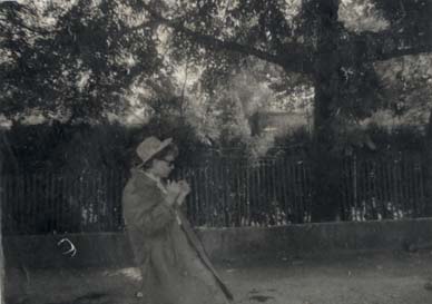 Homme de profil en imperméable et chapeau devant une palissade avec jardin et maison. Noir et blanc.