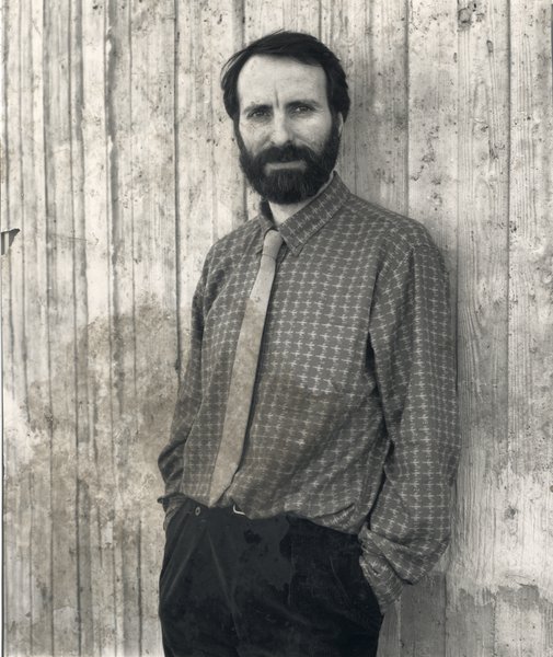 Homme barbu debout en chemise et cravate devant une palissade en bois. Noir et blanc.