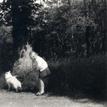 Petite fille blonde en kilt avec un chien en laisse devant buissons et d’arbres. Noir et blanc.