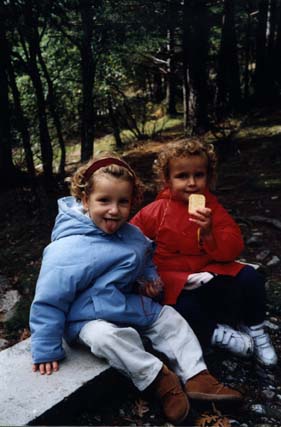 Deux petits enfants assis sur un banc, l’un blond langue tirée en anorak bleu, l’autre un biscuit à la main en anorak rouge, arbres et  végétations en arrière-plan.