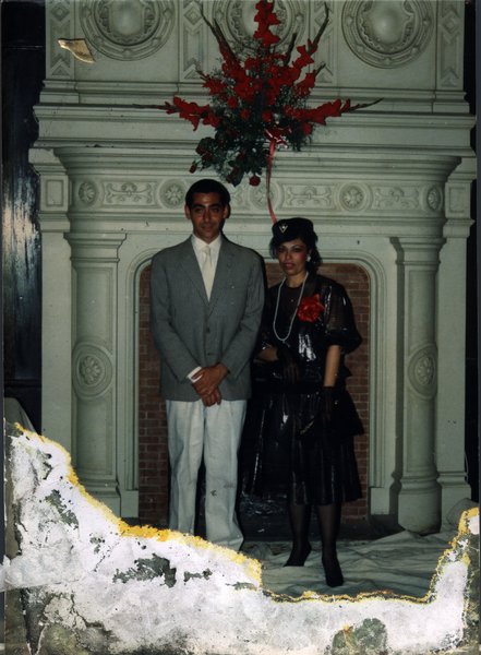 Homme en costume et femme en habits chics et toque noire debout devant une grande cheminée en marbre blanc. Image abîmée