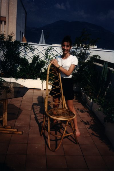 Jeune femme sur une terrasse tenant une chaise en rotin, montagne en arrière-plan.ᴬᴵ