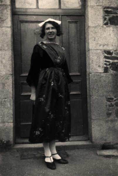 Femme en tenue traditionnelle et coiffe devant une porte. Noir et blanc.ᴬᴵ