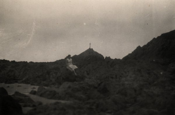 Personne assise sur des rochers face à une croix au sommet d'une colline. Noir et blanc.ᴬᴵ