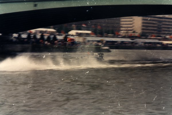 Bateau de course sur une rivière sous un pont, immeubles en arrière-plan.ᴬᴵ