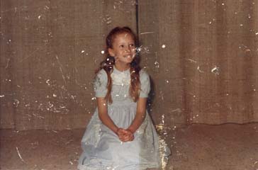 Petite fille blonde en robe à smocks agenouillée devant un rideau.