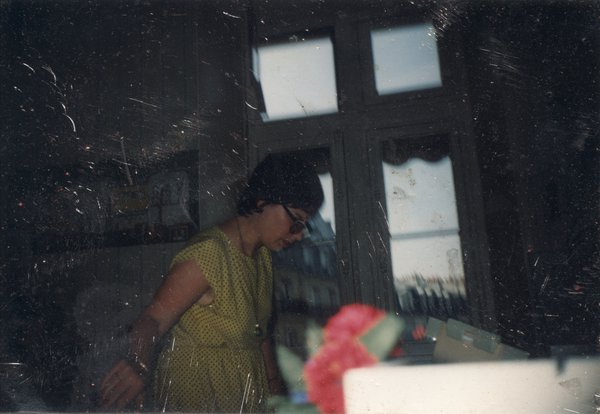 Jeune femme brune à lunettes noire de profil penchée devant une fenêtre, fleurs rouges en avant-plan.