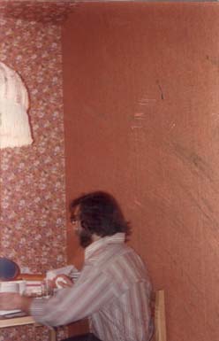Homme barbu à lunettes assis de profil sur une chaise devant un mur tapissé de papier à fleurs.