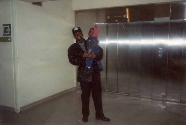 Homme à casquette avec un enfant dans les bras devant les portes d’un ascenseur.